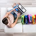 The top view of a woman sitting on a white couch holding a laptop with multi-colored shopping bags next to her.